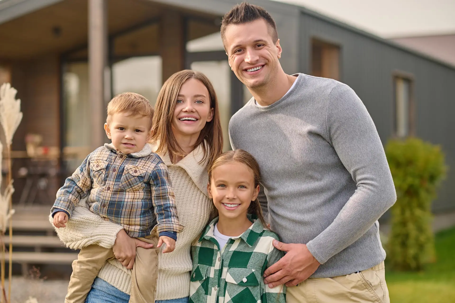 Eine Familie mit zwei Erwachsenen und zwei Kindern steht vor ihrem Haus mit neuer Pergola und schaut in die Kameria
